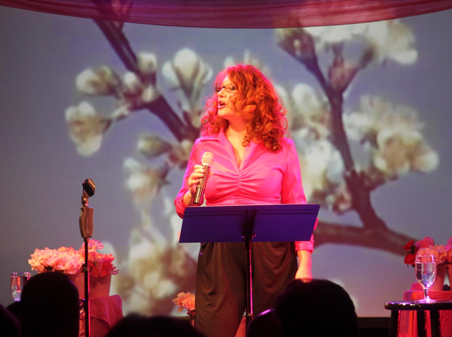 Karen Finley behind a lecturn performing in pink top in front of projection of flowers
